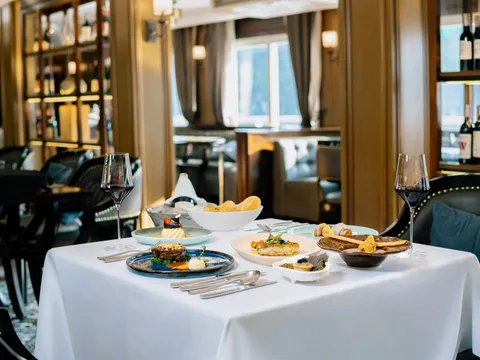 A formal dining table set with multiple plated dishes, wine glasses, and silverware inside an elegant restaurant with wood paneling, wine displays, and windows showing water views.
