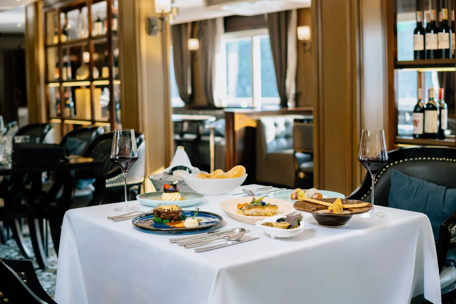 A formal dining table set with multiple plated dishes, wine glasses, and silverware inside an elegant restaurant with wood paneling, wine displays, and windows showing water views.