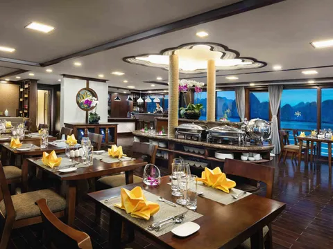A dining room on a cruise boat with set tables, yellow napkins, a buffet station, and large windows showing karst mountains and water at dusk.