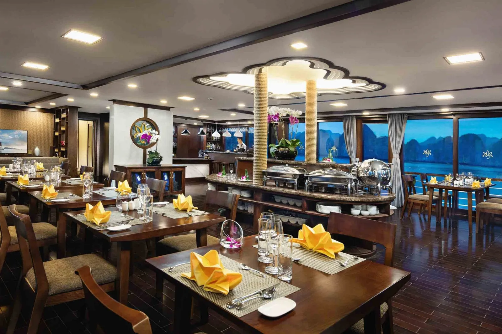 A dining room on a cruise boat with set tables, yellow napkins, a buffet station, and large windows showing karst mountains and water at dusk.