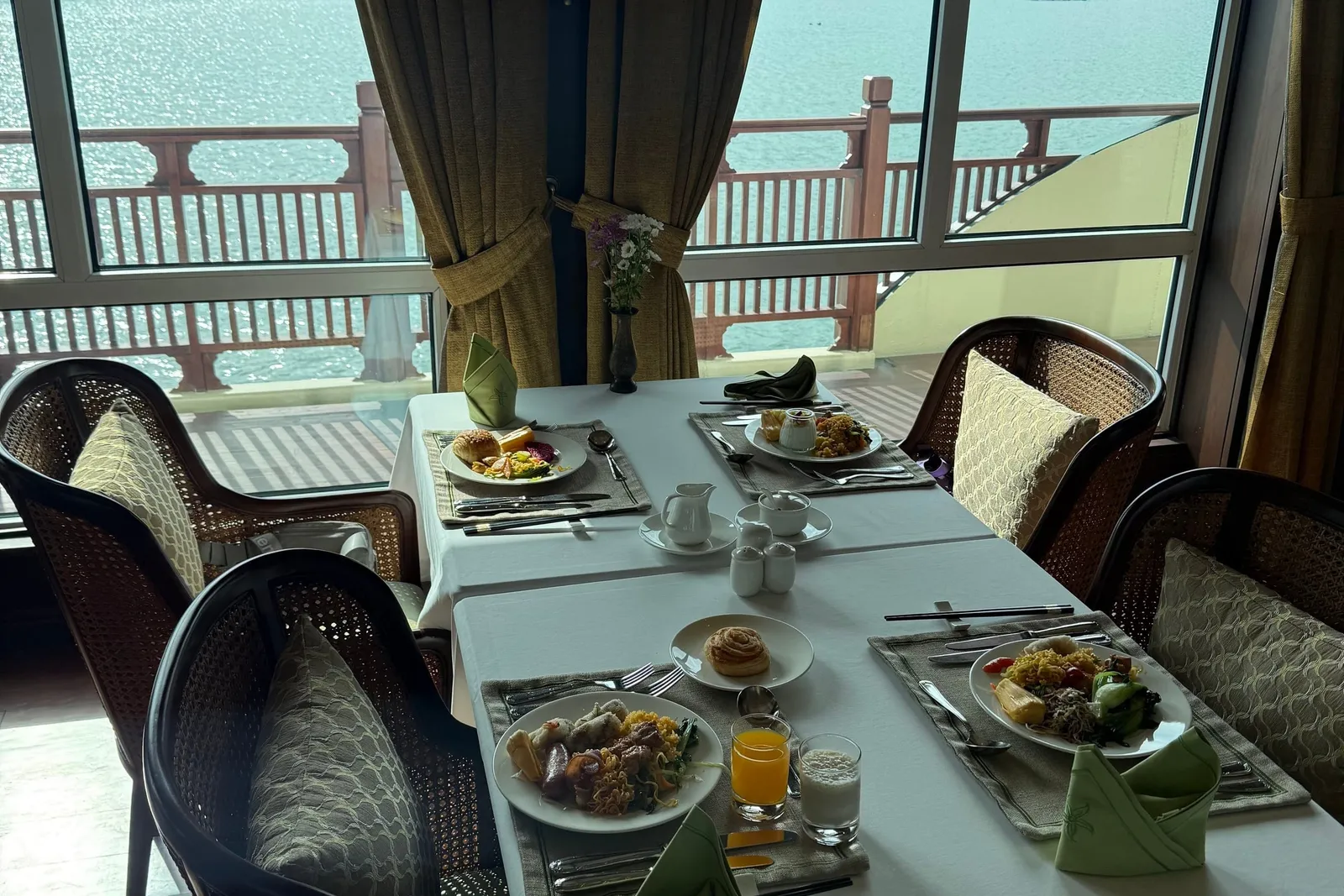 A dining room with white-clothed tables set with breakfast plates, juice and green napkins, beside large windows looking out over a balcony railing to calm water and limestone karst islands.