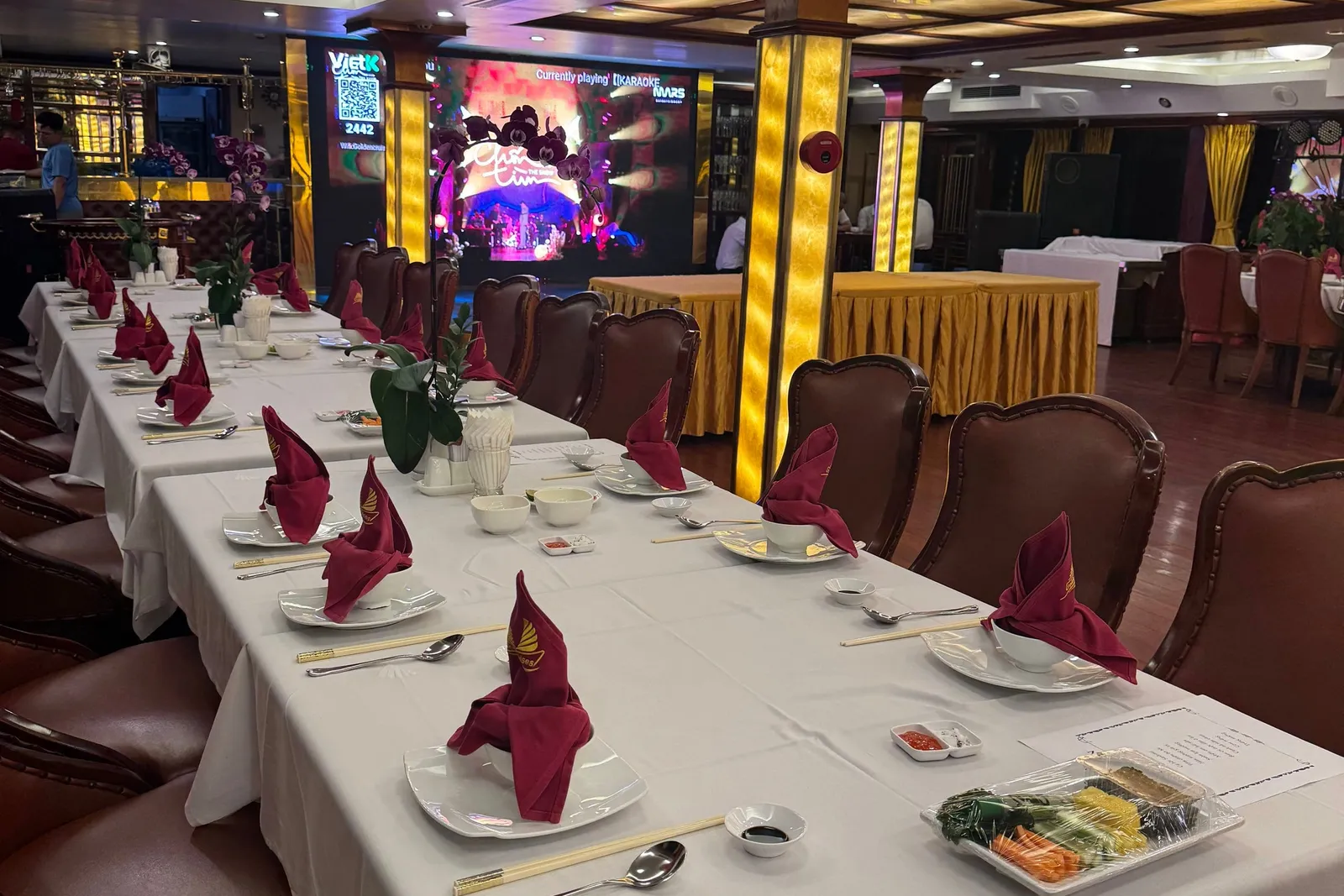 Long table laid for dinner with red napkins folded upright, the stage lit at the far end of the room