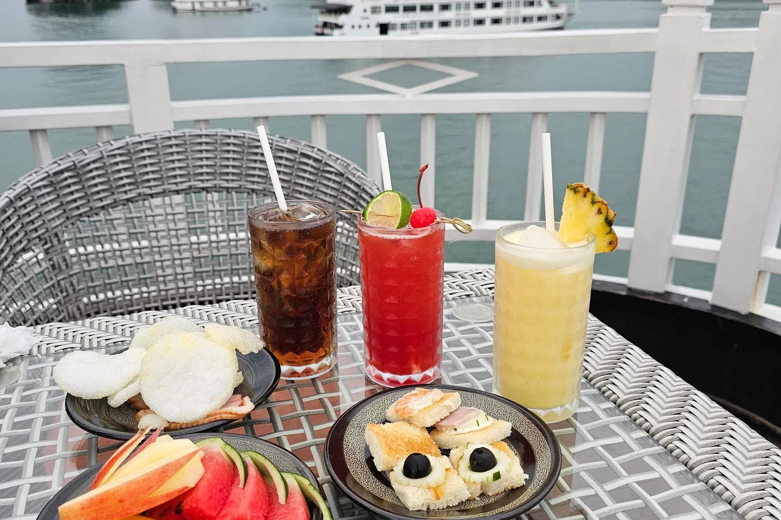 A wicker table on a white-railed deck holds three drinks, plates of fruit, canapes and prawn crackers, with limestone karsts and a white multi-deck cruise ship in the bay behind.