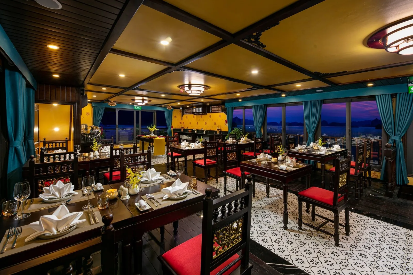 Interior of a ship dining room with dark wooden tables set with folded white napkins, red cushioned chairs, teal curtains and windows showing karst islands at dusk.