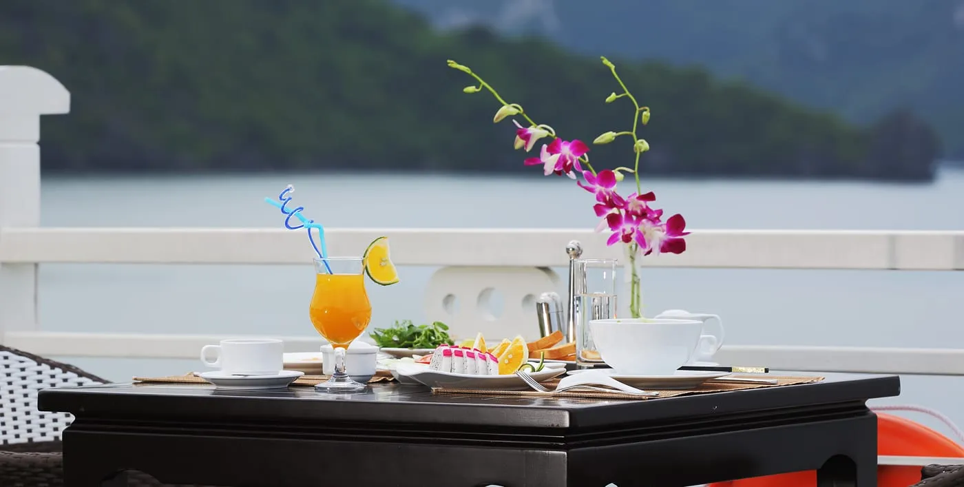 A breakfast table on a white-railed deck holds orange juice, cups, fruit, a salad and a vase of purple orchids, with a calm bay and green forested hills blurred behind and an orange life ring at the lower right.