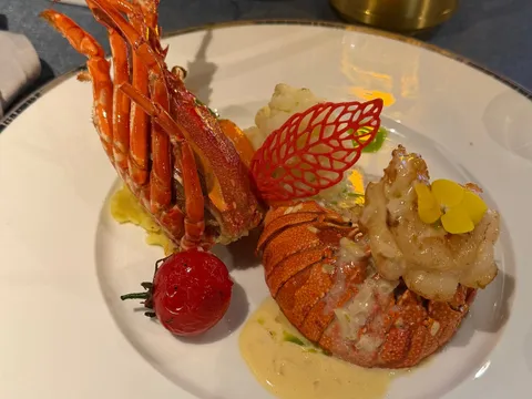 A plated lobster dish with a red tuile leaf, cherry tomato, cauliflower, cream sauce and a yellow flower on a white rimmed plate, set on a dining table with a glowing gold lamp, vase and salt and pepper shakers.