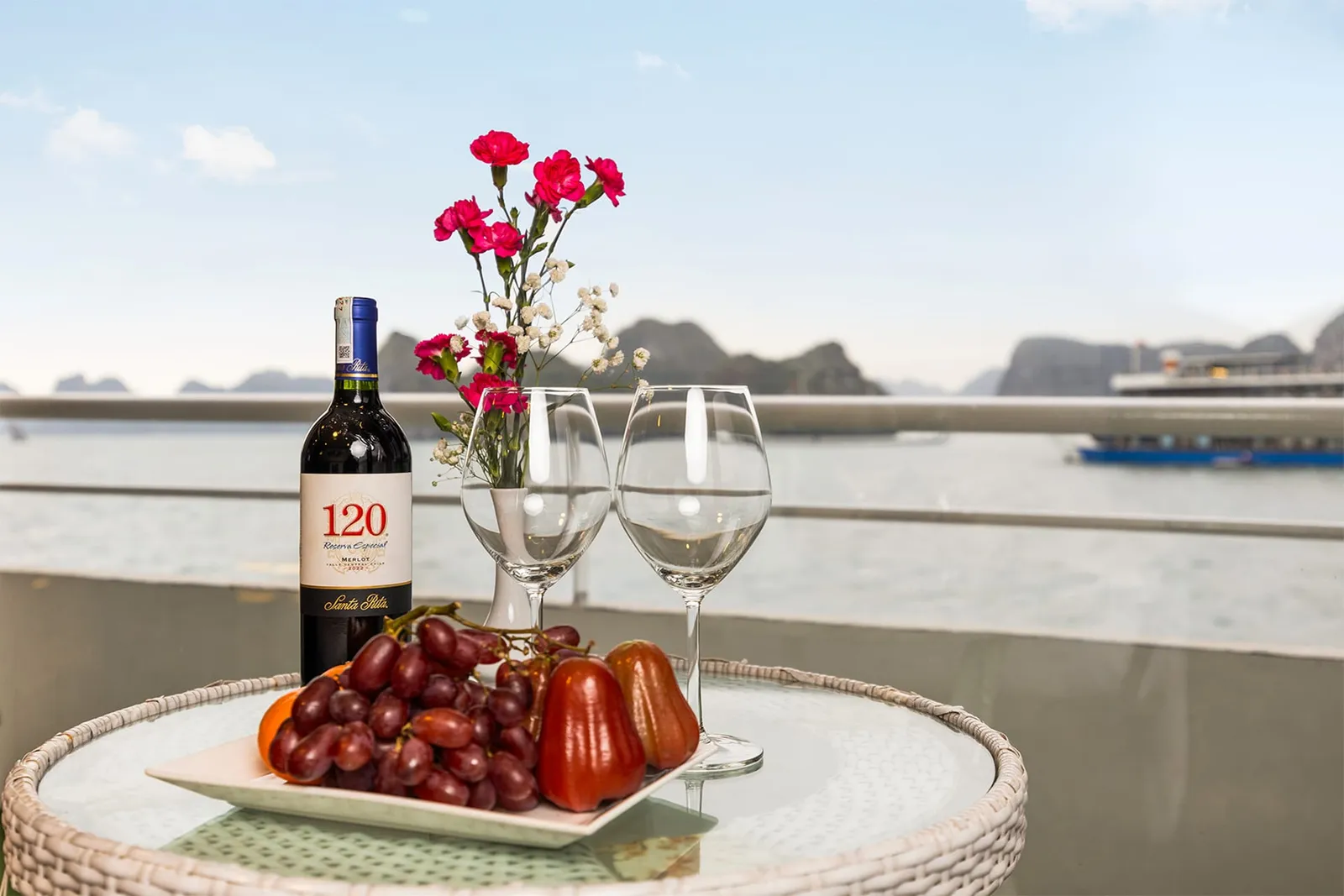 A wicker table on a balcony holds a Santa Rita 120 Merlot bottle, two empty wine glasses, pink carnations in a vase, and a plate of grapes and rose apples, with karst islands and another boat on the water behind.