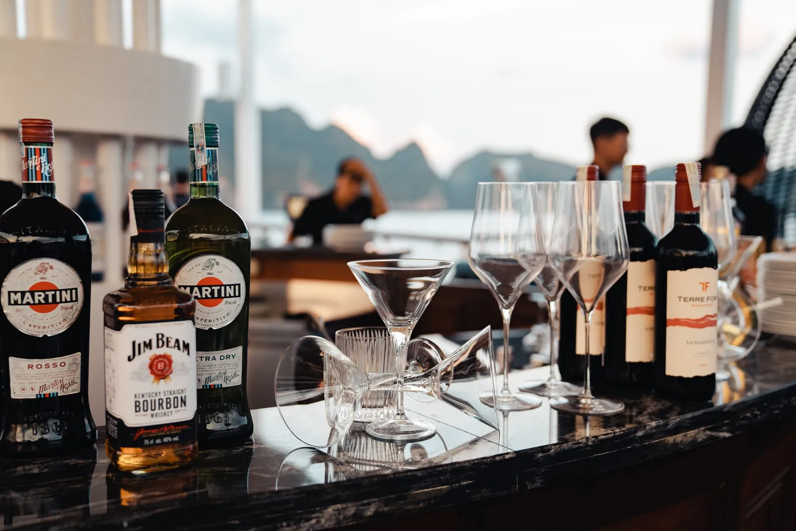 A bar setup with liquor bottles, wine bottles, and glassware on a dark counter with karst limestone peaks visible through windows in the background.