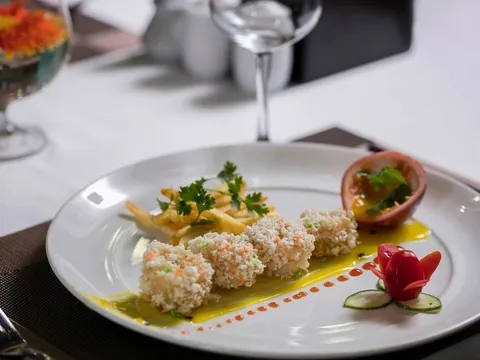 A plated gourmet dish with breaded rolls, garnishes, and decorative elements on a white tablecloth with wine glasses visible in the background.