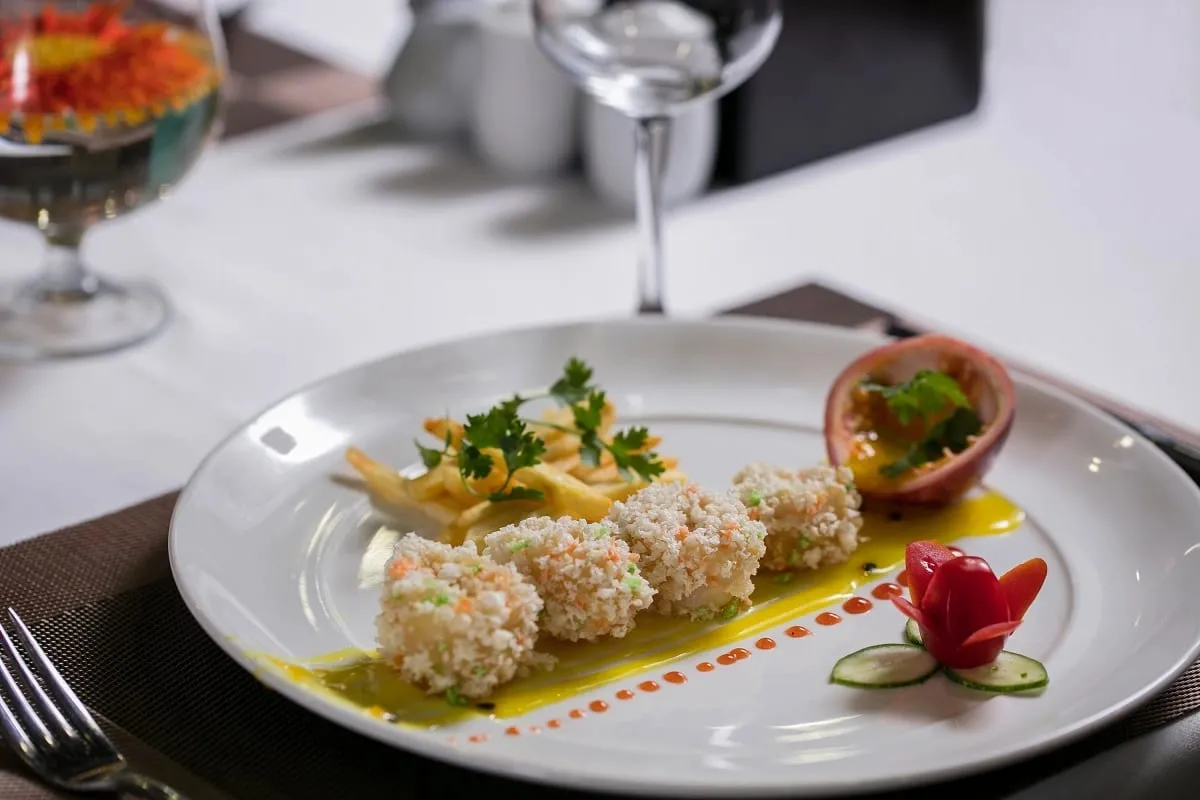 A plated gourmet dish with breaded rolls, garnishes, and decorative elements on a white tablecloth with wine glasses visible in the background.