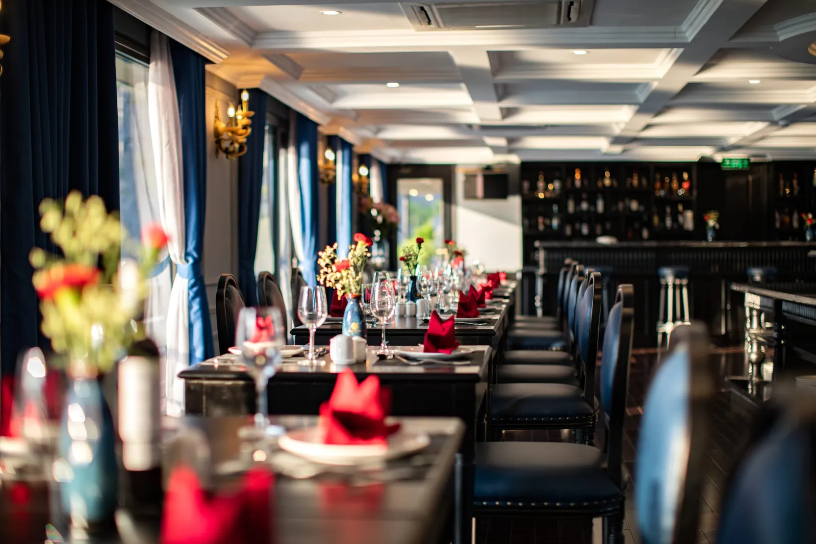 Restaurant laid for dinner: white cloths, red roses on every table, blue drapes between the windows