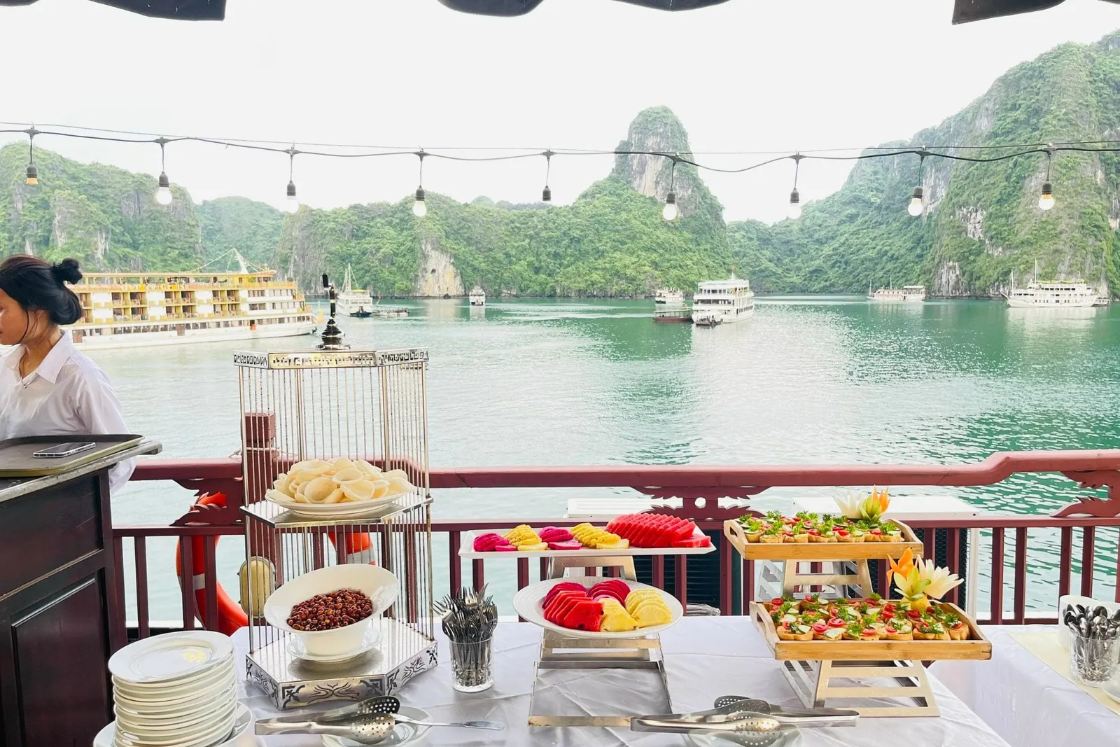 A buffet table with fruit platters, canapés and plates on a boat deck with string lights, a staff member at left, and limestone karsts and other cruise ships on green water beyond the red railing.