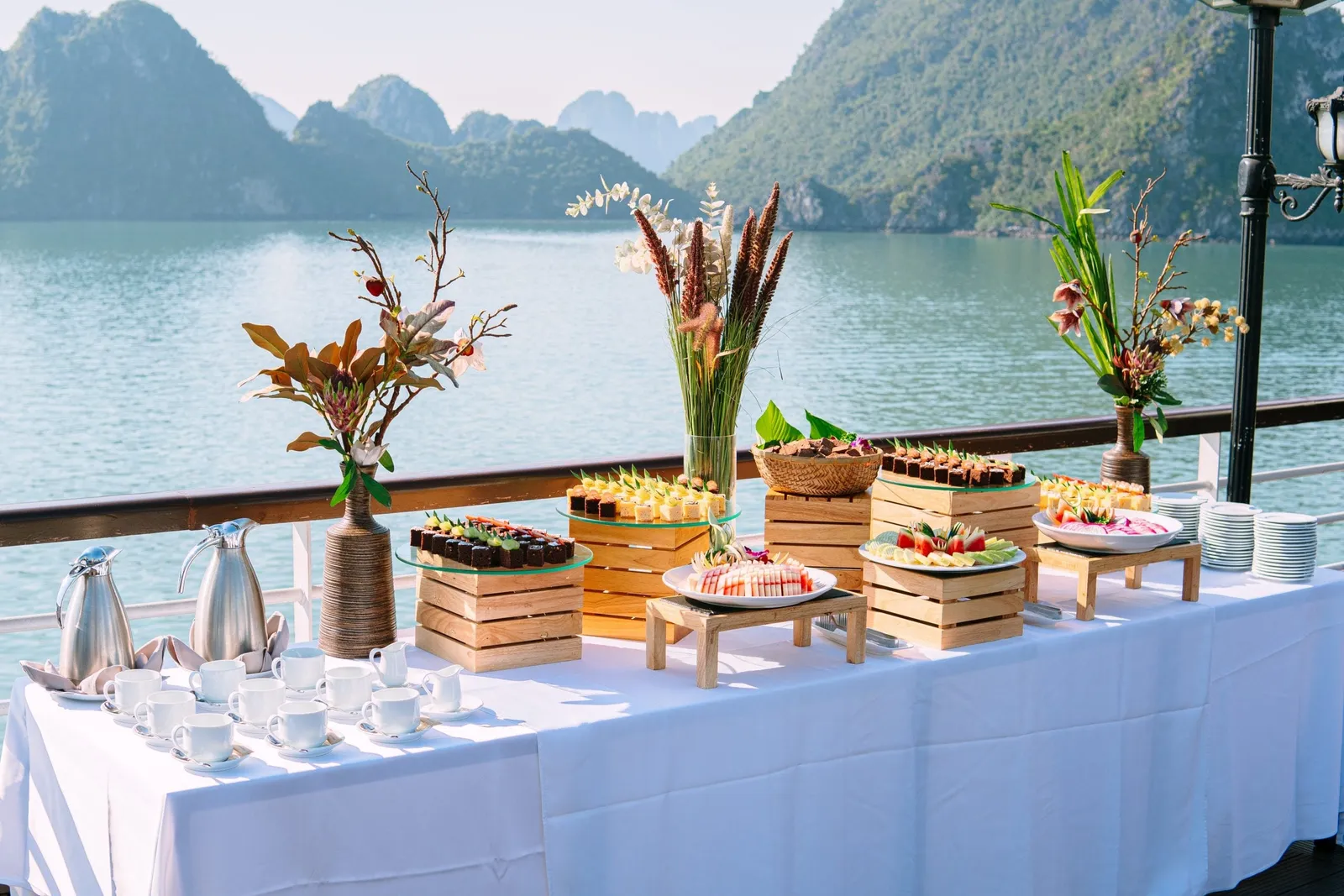 A white-clothed buffet table on a deck with desserts, fruit, coffee cups, flower arrangements and steel pitchers, overlooking calm water and limestone karst islands.