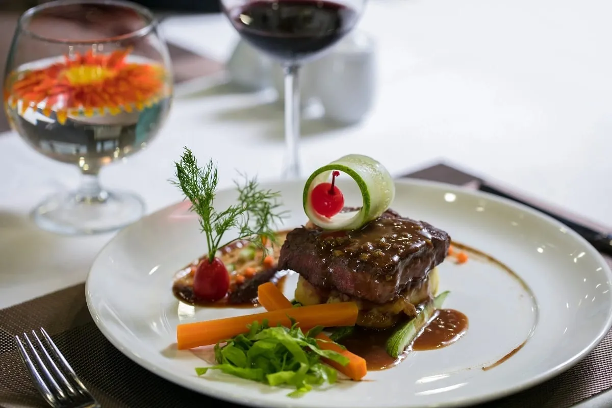 A plated steak dish with vegetables and garnish sits on a white tablecloth with wine glasses in soft focus behind it.