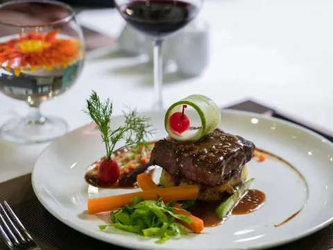 A plated steak dish with vegetables and garnish sits on a white tablecloth with wine glasses in soft focus behind it.