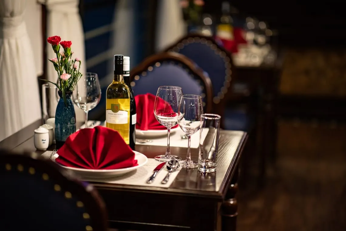 A dark wooden dining table set with red folded napkins, wine glasses, a white wine bottle, a vase of pink and red carnations and blue studded chairs in a dimly lit dining room, with small branding on the salt and pepper shakers and bottle label.