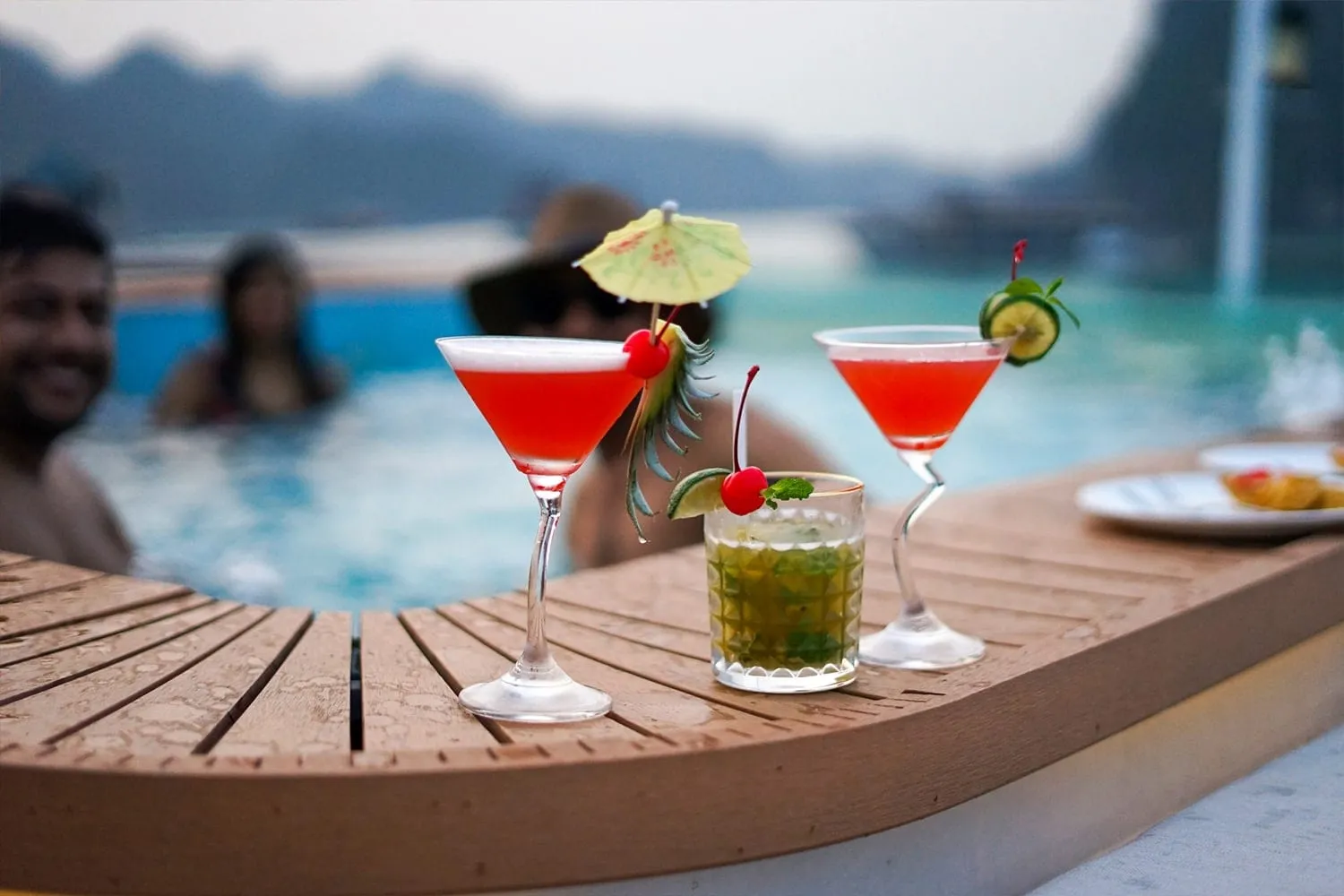 Two red cocktails and a mojito on a wooden pool-side bar with blurred people swimming in a pool and hazy karst hills in the background.