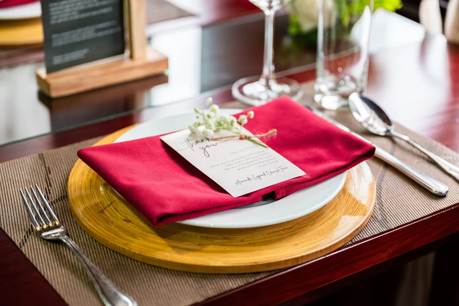 A dining table place setting with a red folded napkin on a white plate and bamboo charger, topped with a printed thank-you card signed by a cruise team, plus fork, spoon, wine glasses and a menu stand.