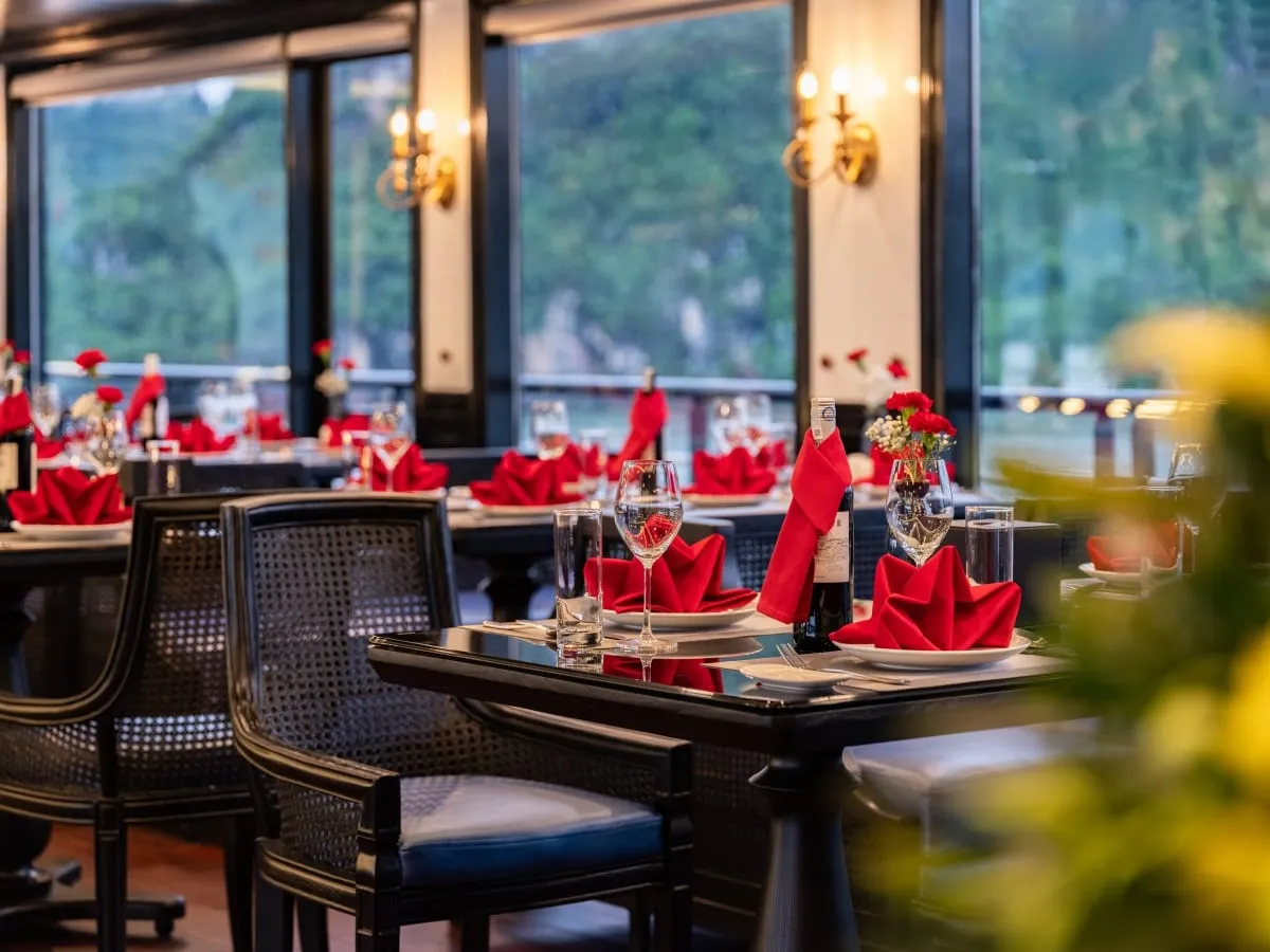 A dining room with black tables set with red folded napkins, wine glasses and small flower vases, with wall sconces and large windows showing green forested hills outside.