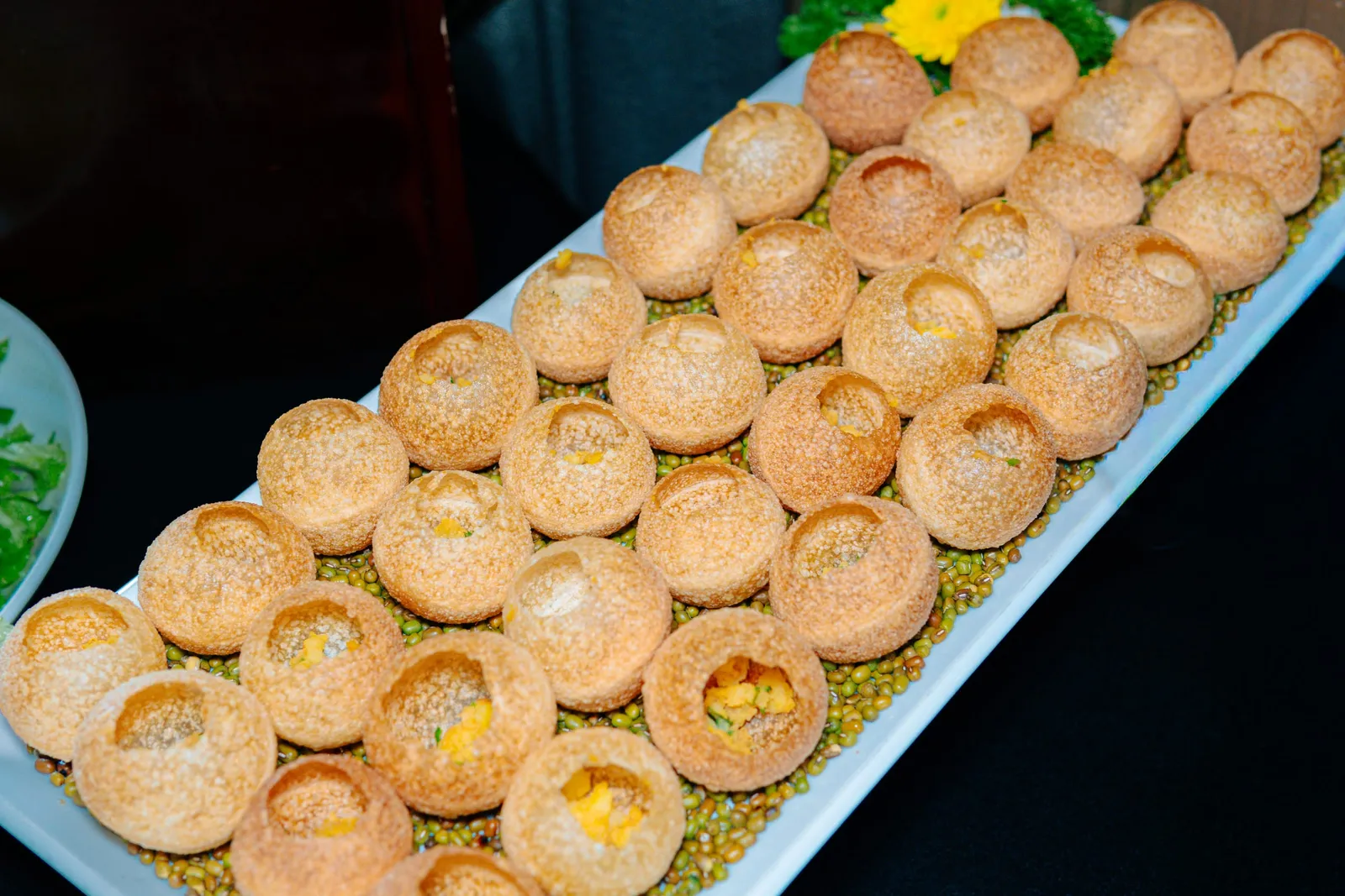 A white rectangular platter displays rows of round golden pastries with hollow centers filled with yellow filling, arranged on a bed of green beans and corn.