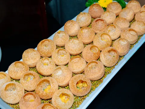 A white rectangular platter displays rows of round golden pastries with hollow centers filled with yellow filling, arranged on a bed of green beans and corn.