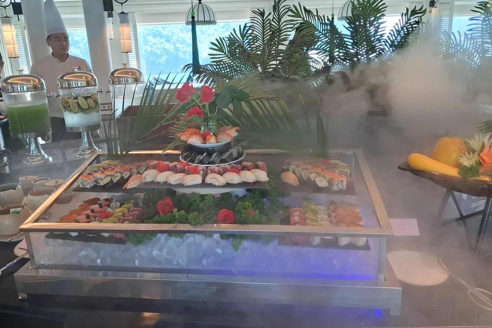 A sushi buffet display on ice with dry-ice fog, flowers and palm fronds, drink dispensers, and a chef in a tall white hat standing behind it in a bright dining room with large windows.