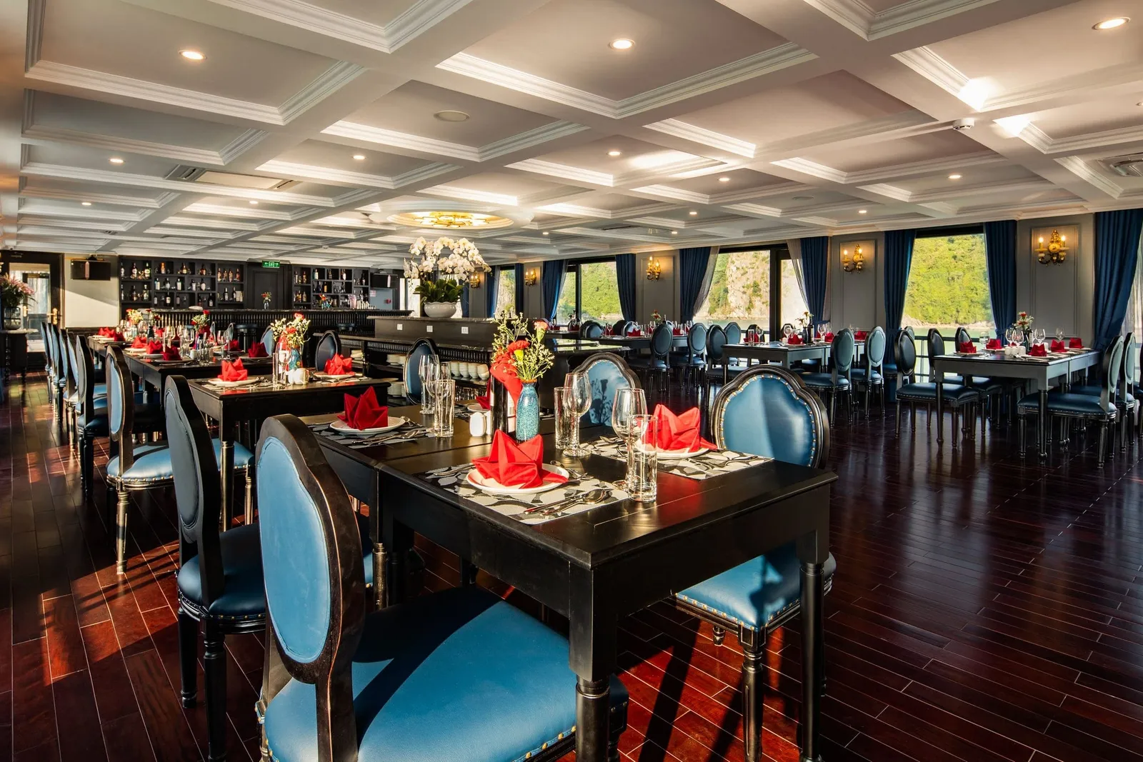 An elegant dining room with dark wood tables set with red napkins, blue upholstered chairs, coffered ceiling, chandelier, bar area, and windows showing green karst formations outside.
