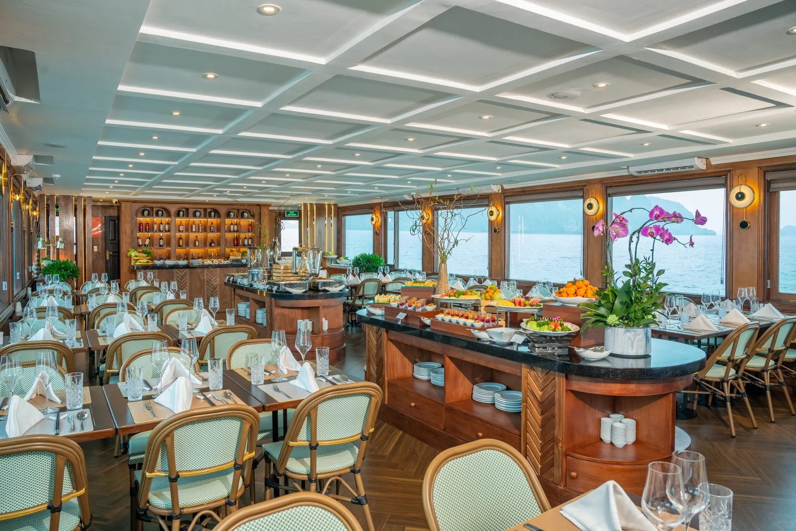 Interior of a ship restaurant with set tables, green rattan chairs, buffet islands with fruit, and large windows showing the sea and karst islands.