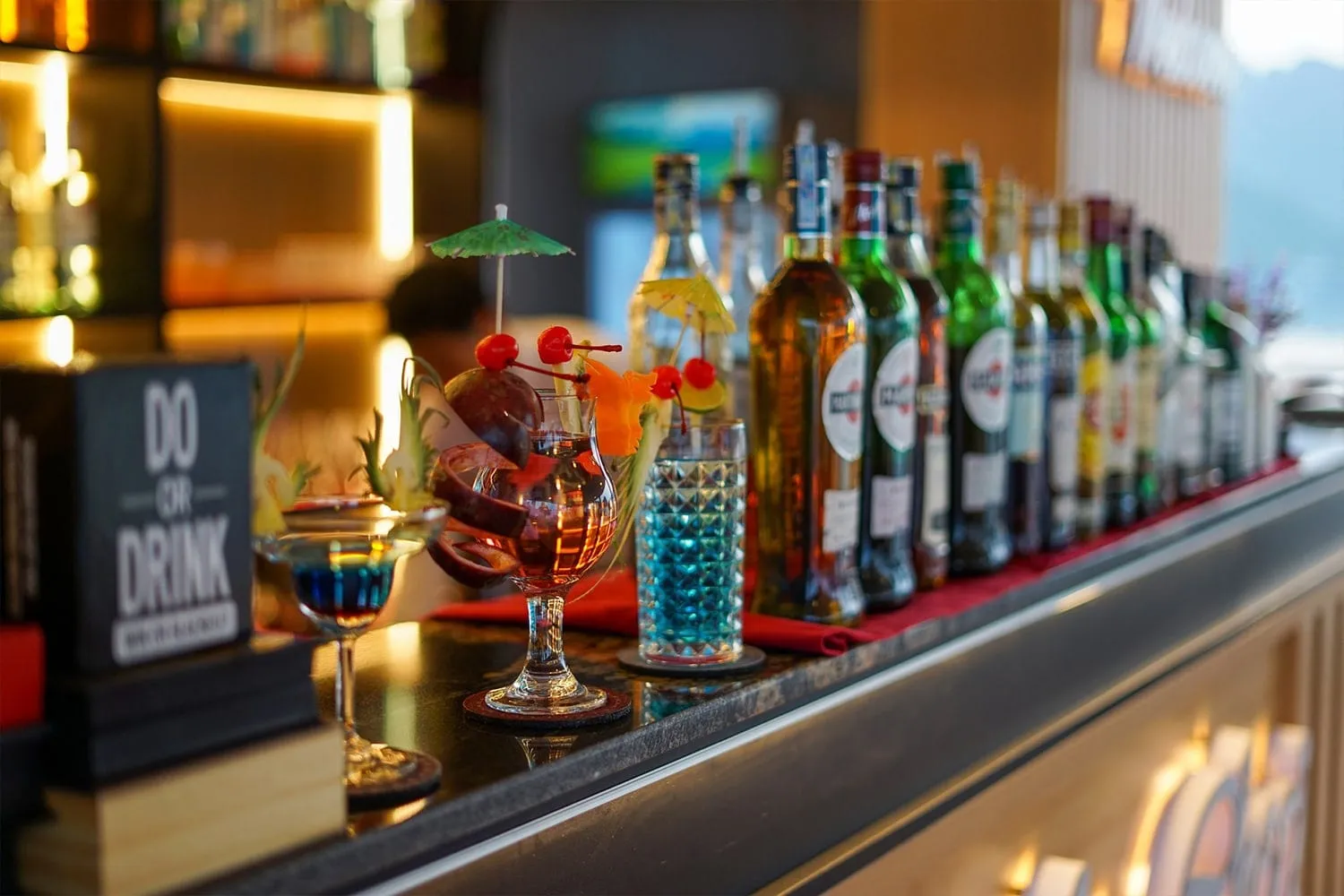A bar counter with garnished cocktails and a row of liquor bottles, a 'Do or Drink' sign on the left, and blurred misty karst hills visible at the upper right.