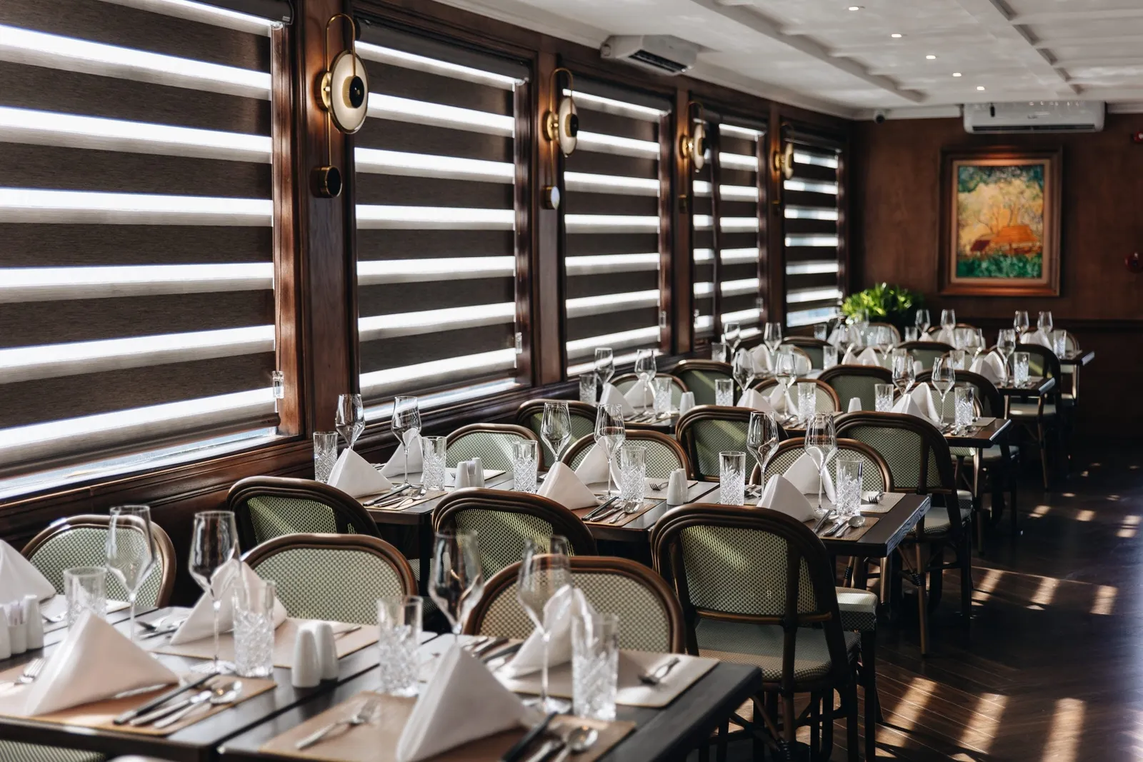 An indoor dining room with rows of tables set with glasses, folded napkins and cutlery, rattan-backed chairs, dark wood paneling and window blinds letting in sunlight.