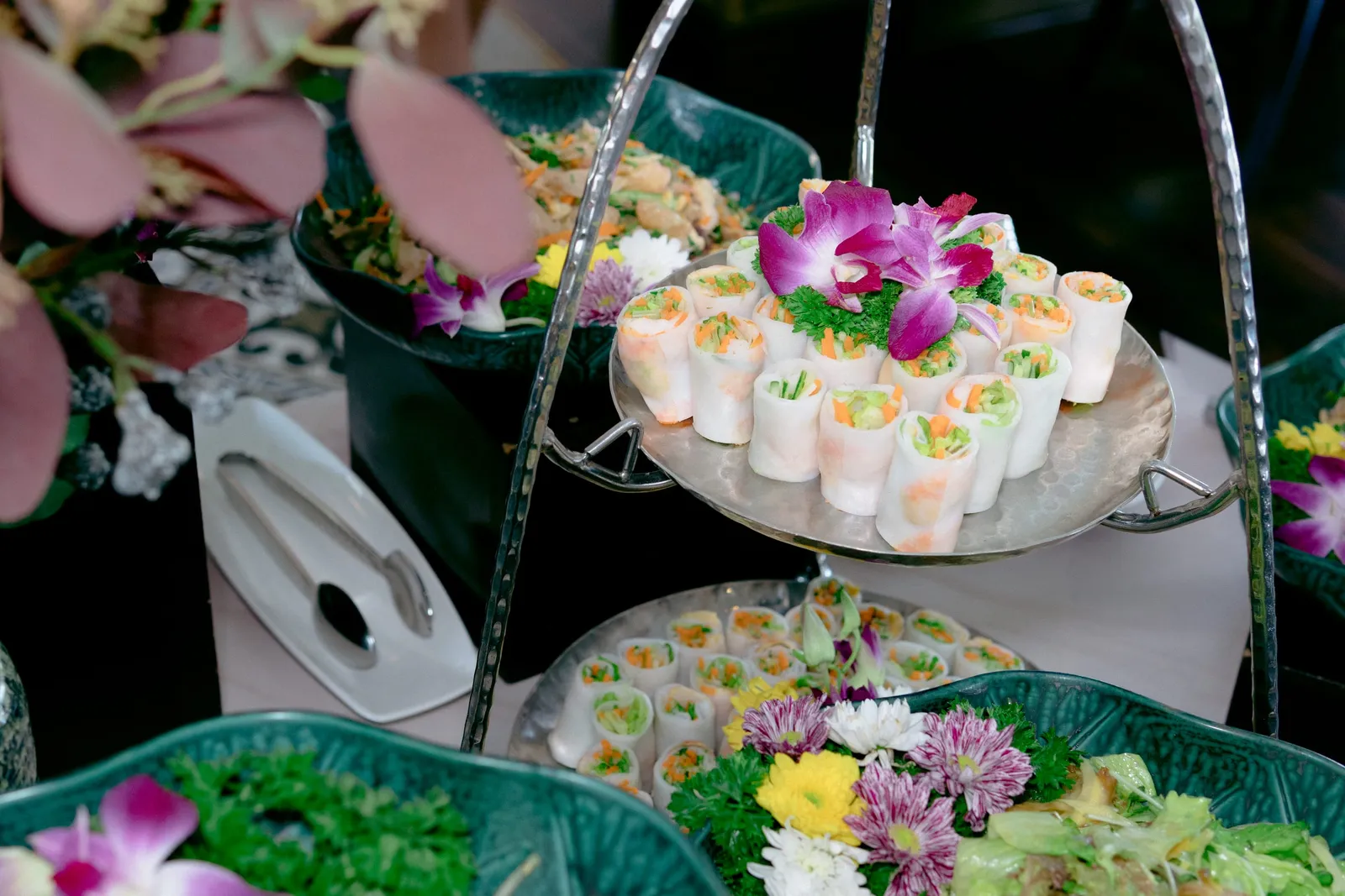 A tiered serving stand displays spring rolls and salads garnished with orchids and chrysanthemums.