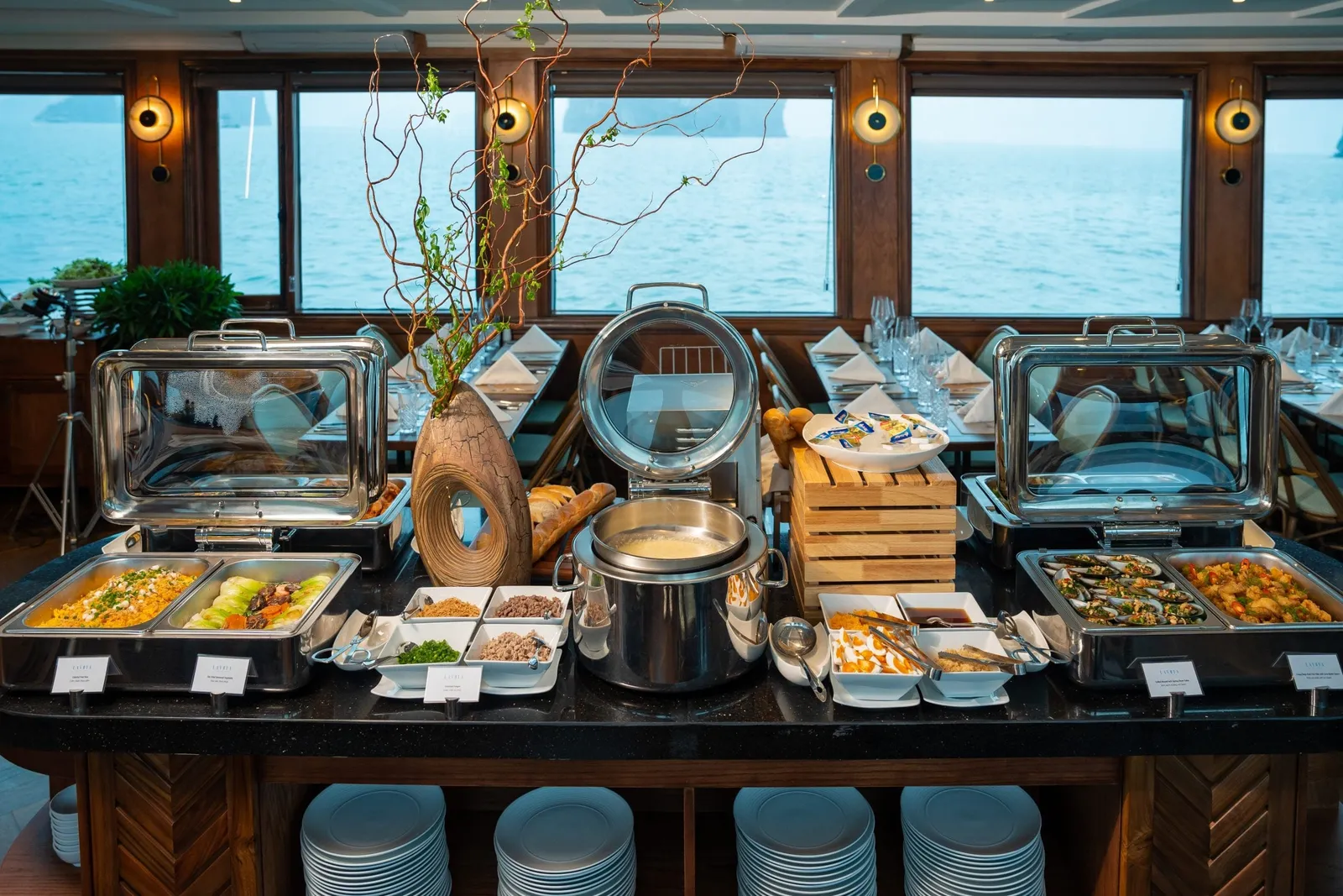A buffet counter with chafing dishes, a pot, bread and small bowls in a dining room with set tables, with windows looking out to calm sea and distant karst islands.