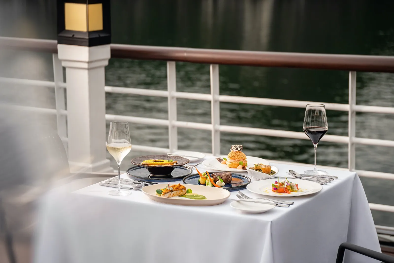 A white-clothed table on a ship's deck set with several plated gourmet dishes, a white wine glass and a red wine glass, with a white railing and dark water behind.