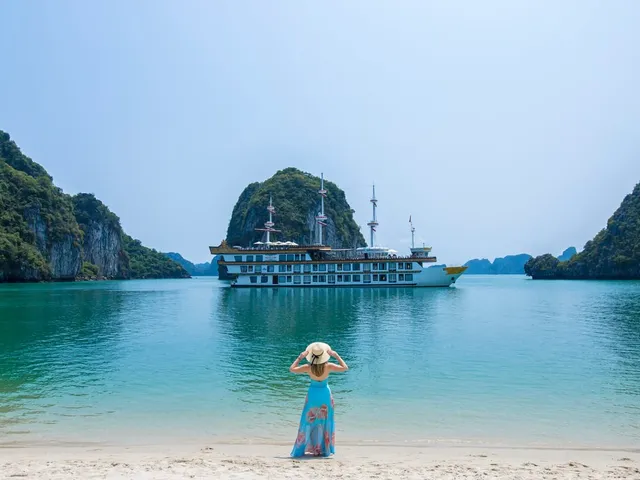 Dragon Legend at anchor off a curve of white sand, a guest in a straw hat standing in the shallows looking back at the ship