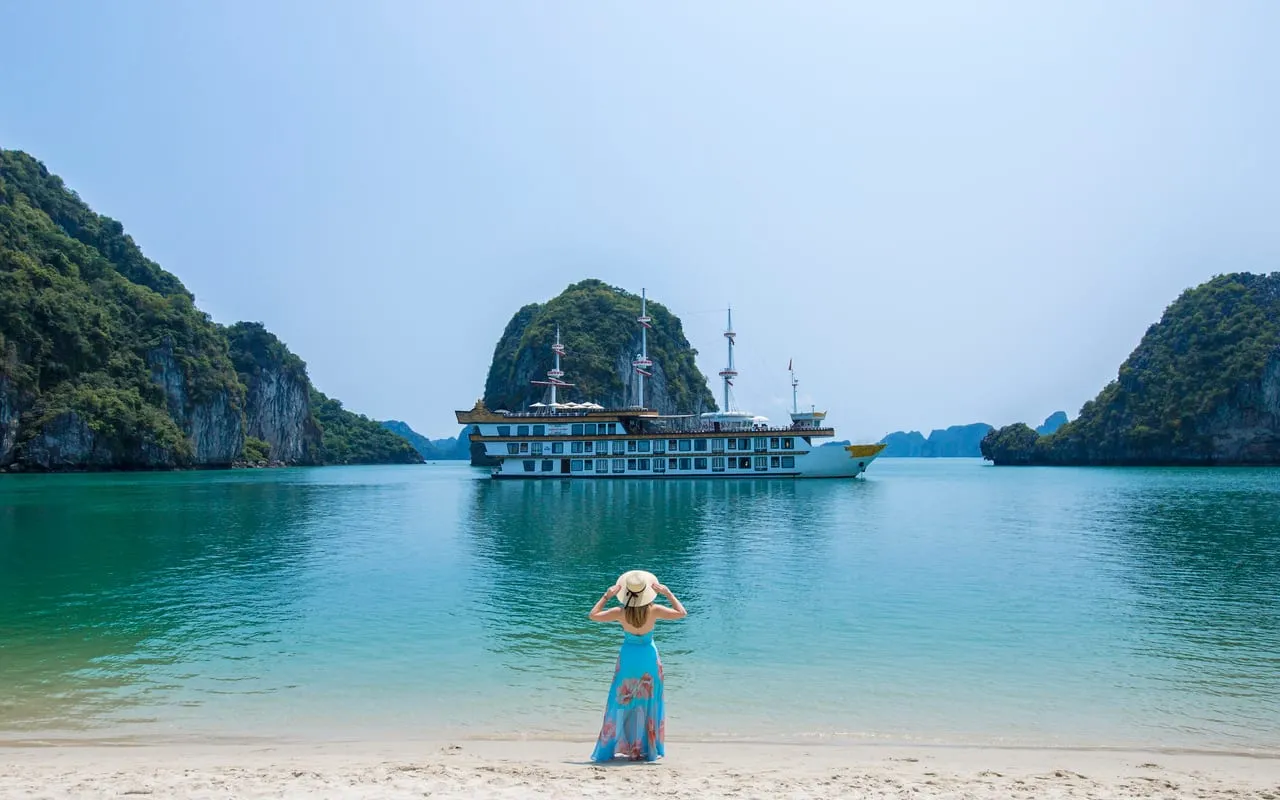 Dragon Legend at anchor off a curve of white sand, a guest in a straw hat standing in the shallows looking back at the ship
