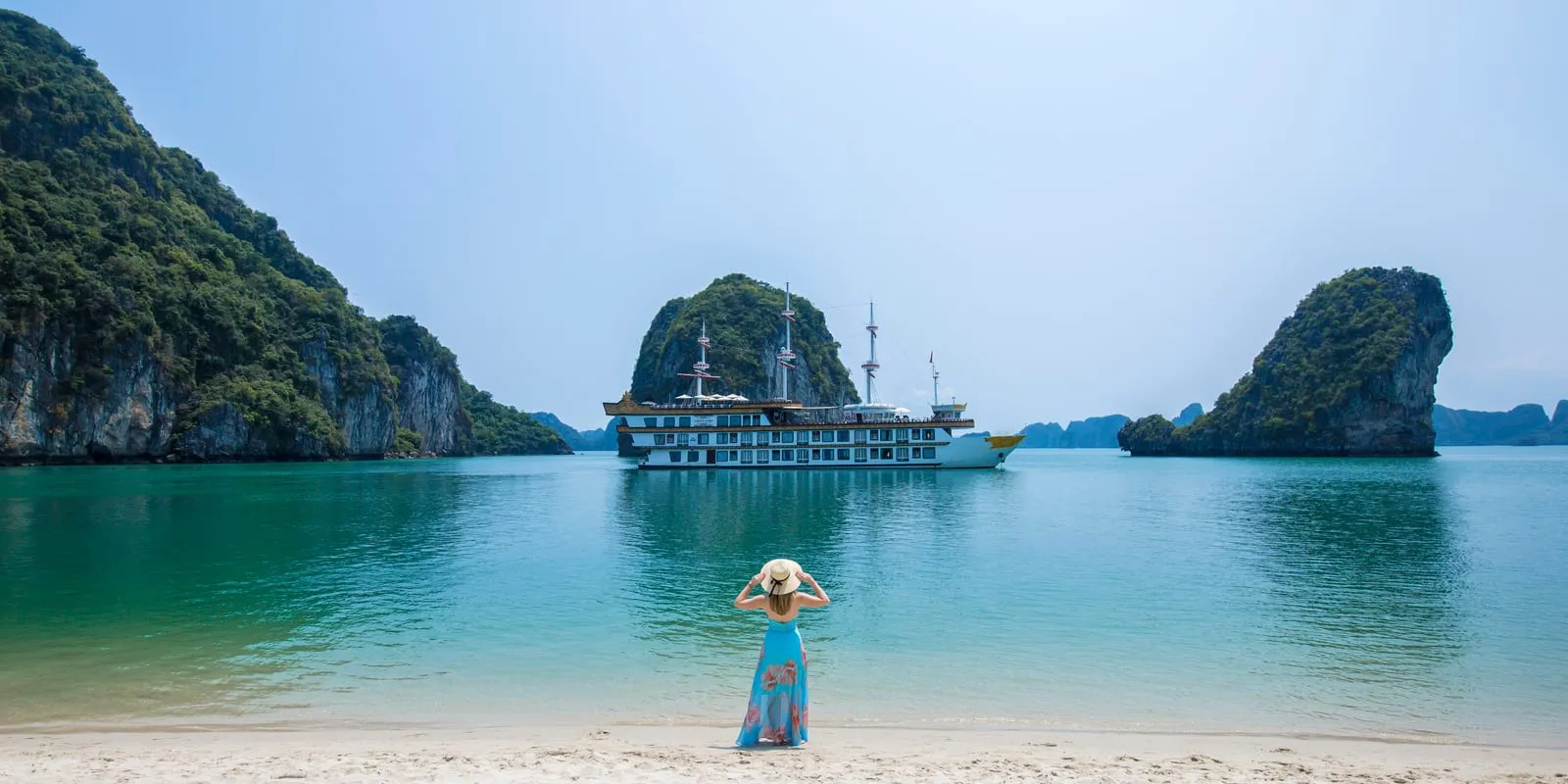 Dragon Legend at anchor off a curve of white sand, a guest in a straw hat standing in the shallows looking back at the ship