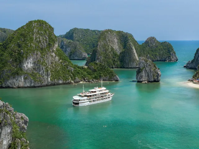A white multi-deck cruise ship anchored in turquoise water surrounded by steep limestone karst islands with a small beach visible on the right.