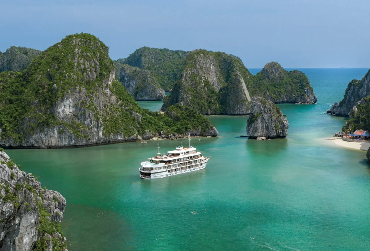 A white multi-deck cruise ship anchored in turquoise water surrounded by steep limestone karst islands with a small beach visible on the right.