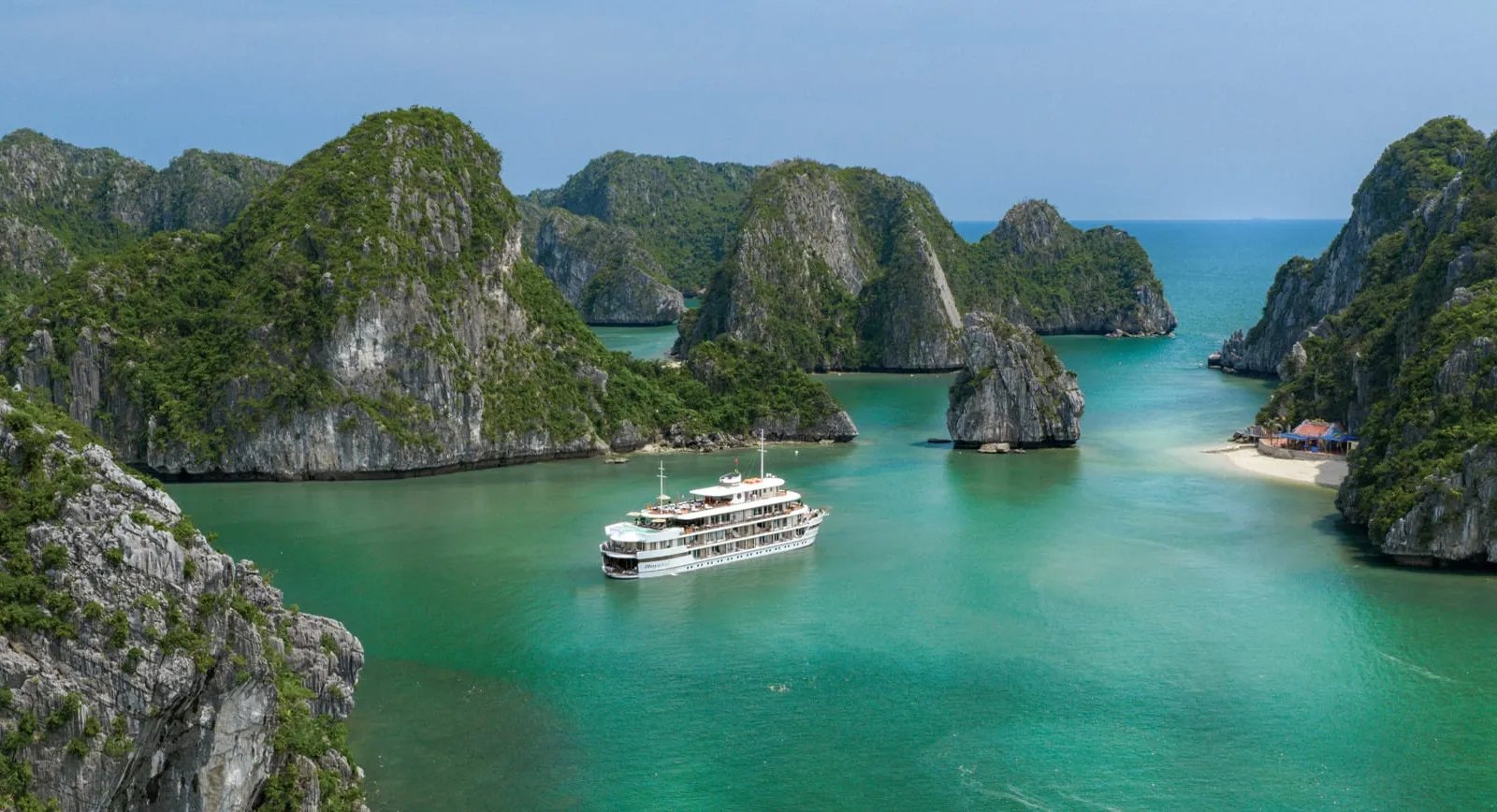 A white multi-deck cruise ship anchored in turquoise water surrounded by steep limestone karst islands with a small beach visible on the right.