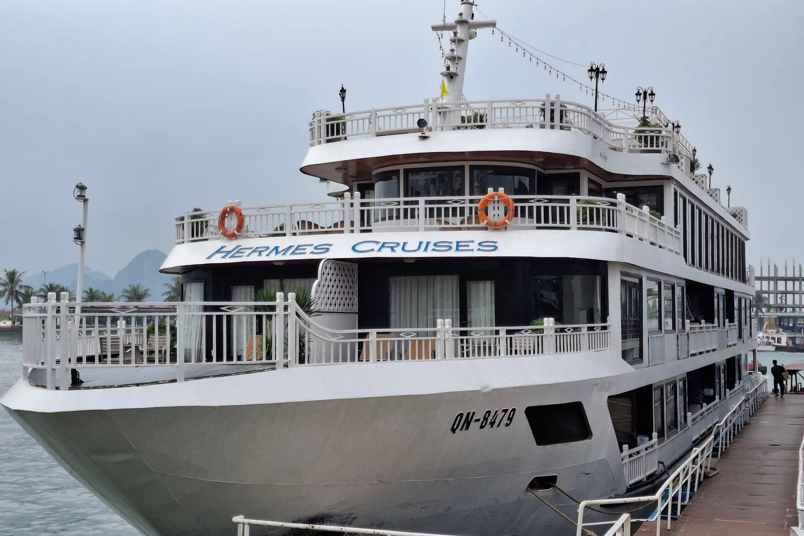 A white multi-deck steel cruise ship moored at a pontoon dock, with 'Hermes Cruises' painted on the upper hull and QN-8479 on the bow, under a hazy sky with karst hills in the distance.