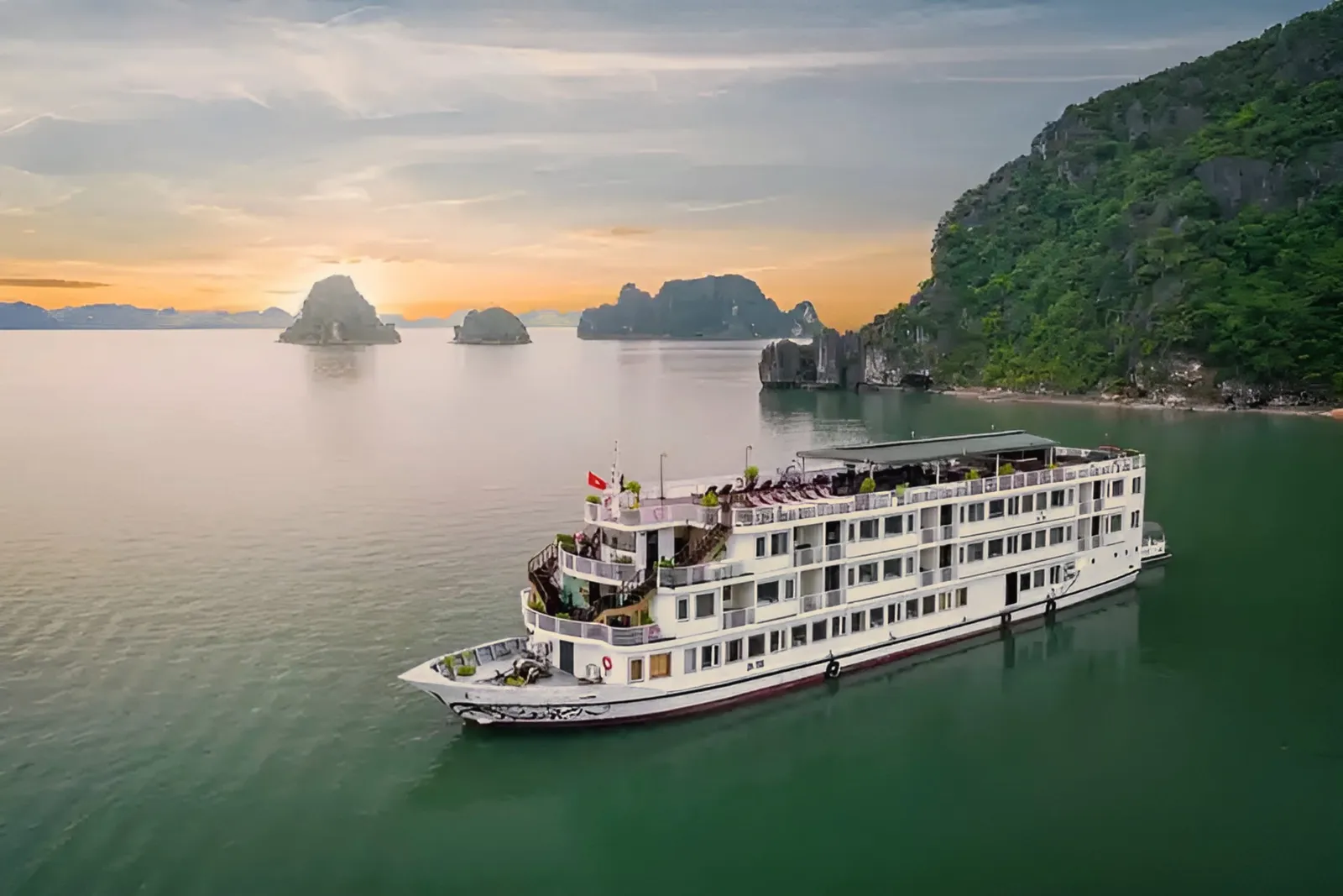 A white multi-deck cruise ship floats in calm green water with limestone karst islands in the background at sunset.