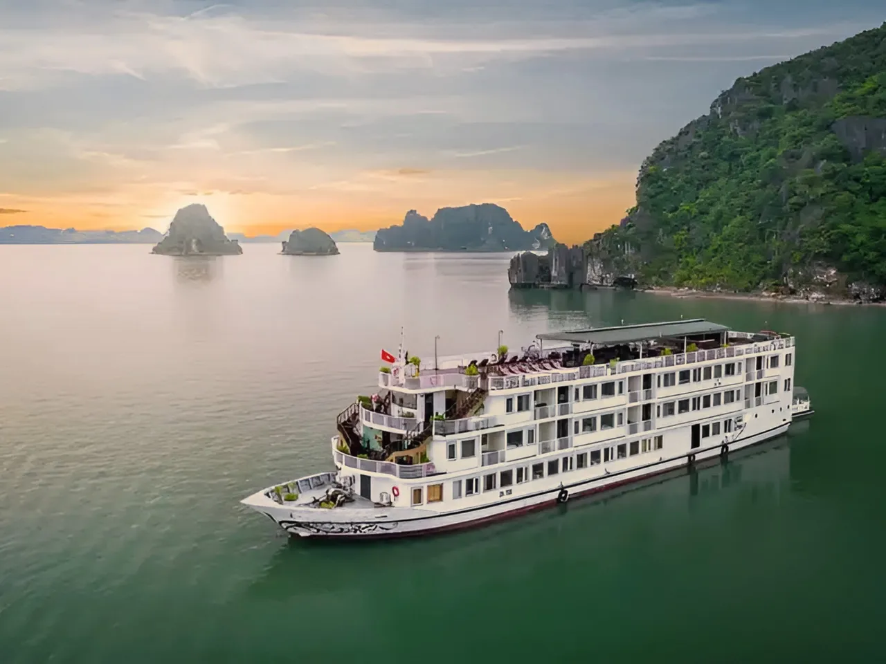 A white multi-deck cruise ship floats in calm green water with limestone karst islands in the background at sunset.