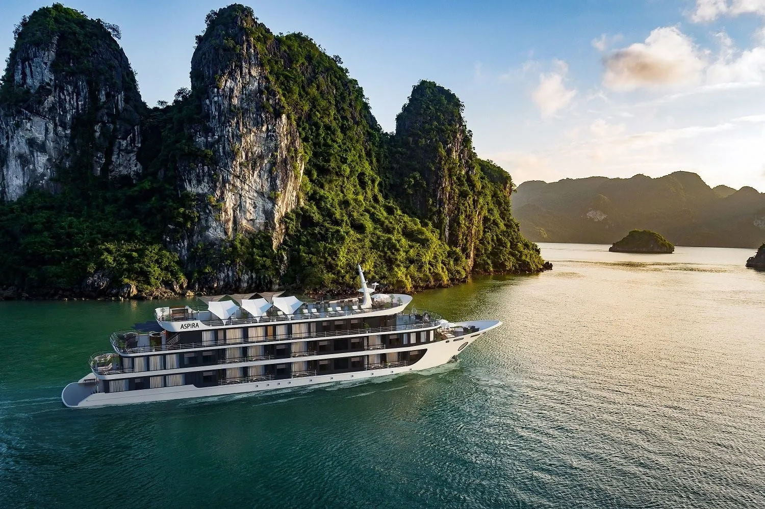 A multi-deck cruise ship with ASPIRA written on its hull sails through green water surrounded by tall limestone karst formations.