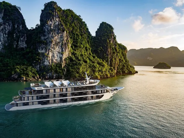 A multi-deck cruise ship with ASPIRA written on its hull sails through green water surrounded by tall limestone karst formations.