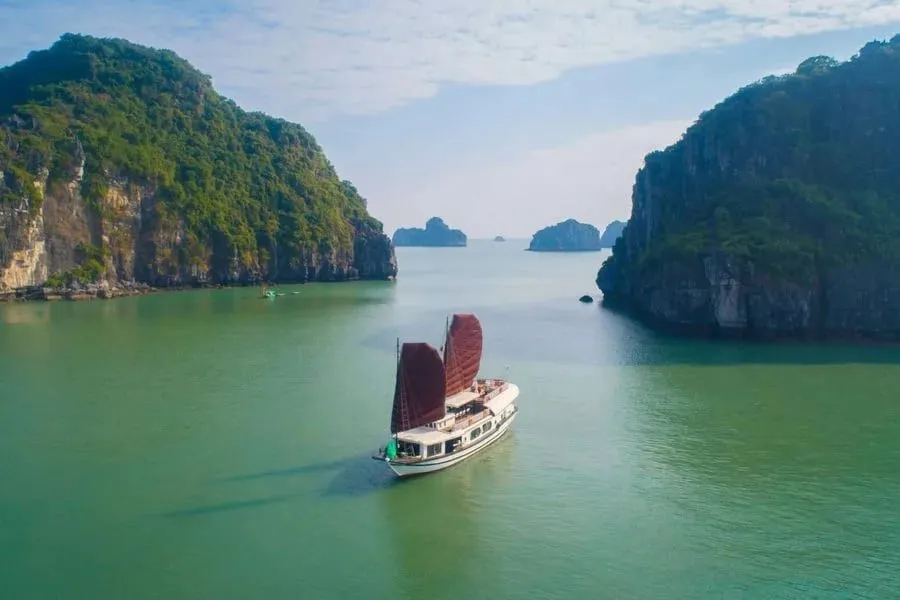 An aerial view of a white junk-style boat with two dark red sails on green water between forested limestone karst cliffs, with more karsts in the hazy distance.