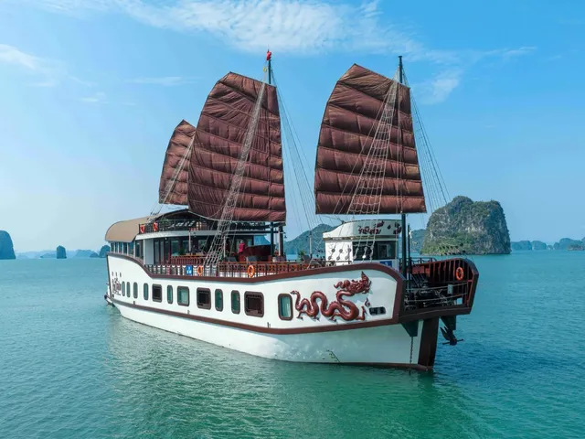 A white junk-style cruise boat with brown sails and a red dragon design on the hull sits on calm green water in front of limestone karst islands under a blue sky.