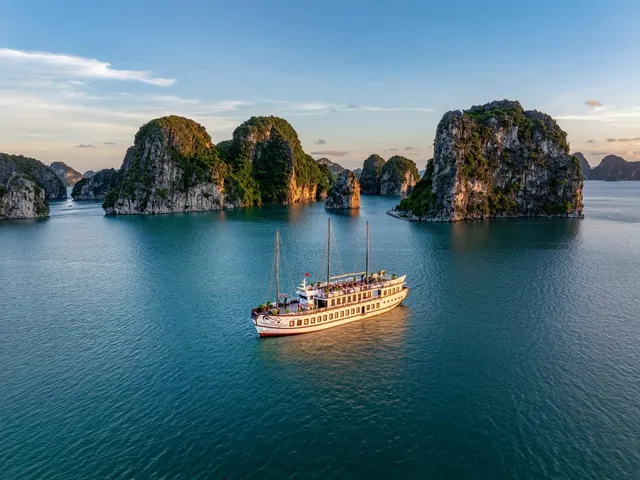 A multi-deck white cruise junk with red trim and a name painted on its hull floats on calm turquoise water surrounded by the iconic karst limestone islands of Ha Long Bay at golden hour.