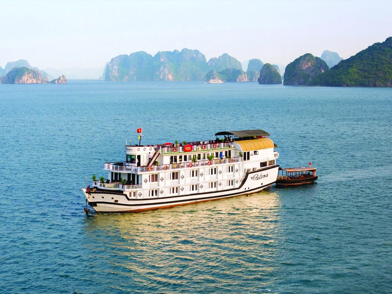 A white and yellow cruise boat with multiple decks floats in calm blue water with distinctive limestone karst mountains in the background.