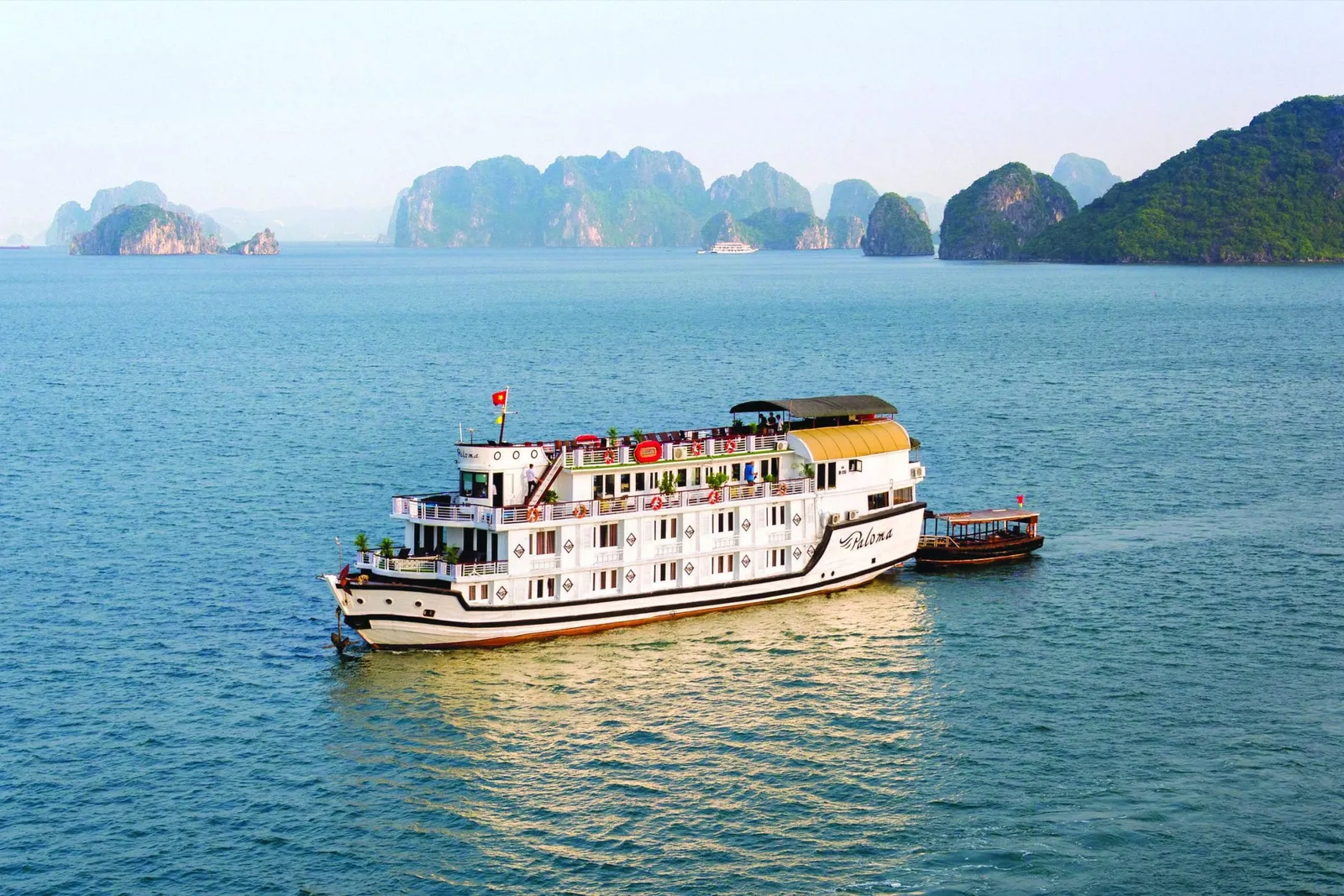 A white and yellow cruise boat with multiple decks floats in calm blue water with distinctive limestone karst mountains in the background.
