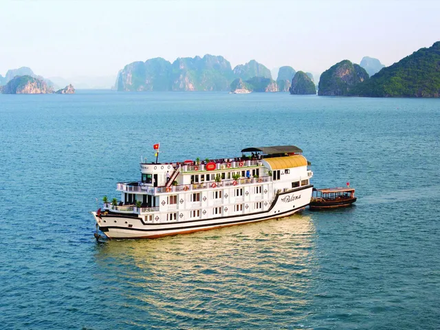 A white and yellow cruise boat with multiple decks floats in calm blue water with distinctive limestone karst mountains in the background.